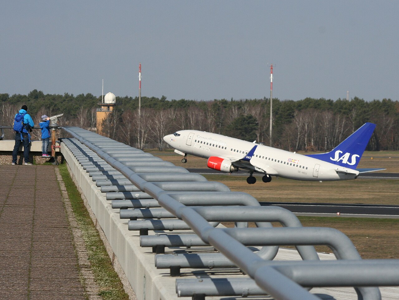 An SAS Boeing 737 taking off from Berlin Tegel, watched by two people on the viewing terrace